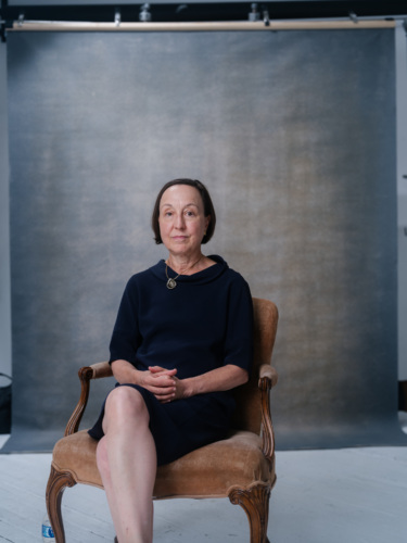 A woman with short brown hair wearing a dark blue dress and a pendant necklace, sitting in an upholstered chair against a grey backdrop.