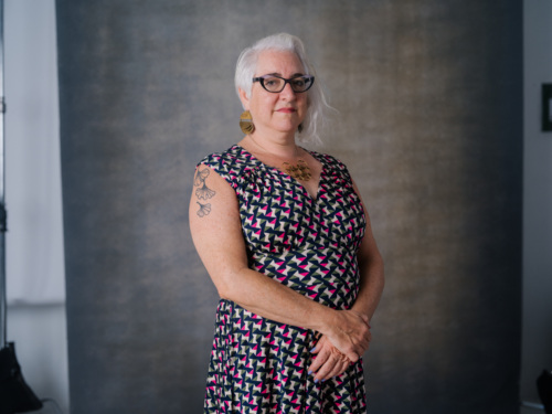 A woman with grey hair and glasses, wearing a geometric print dress and gold statement jewelry, stands with her hands clasped.