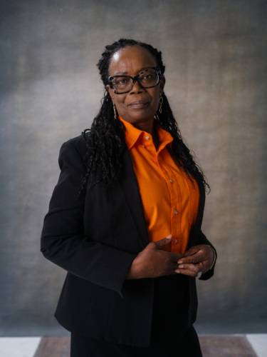 A woman with braided hair and glasses wearing a black blazer over an orange button-down shirt.