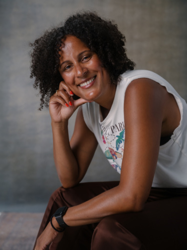 A smiling woman with curly hair, wearing a sleeveless white top, resting her chin on her hand.