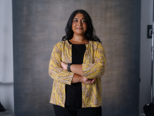A woman with long dark hair stands with her arms crossed, wearing a black shirt under a yellow cardigan patterned with giraffes.