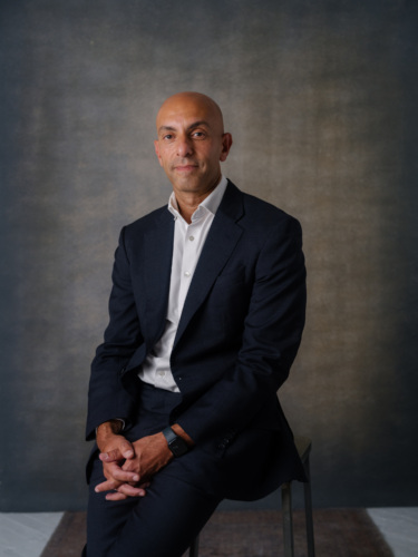 A bald man in a dark suit and white button-down shirt sits on a stool against a mottled gray backdrop.