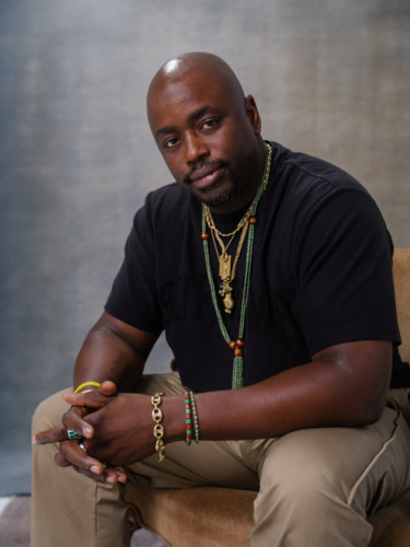 A bald Black man in a black T-shirt and tan pants sitting with his hands clasped, wearing layered necklaces and beaded bracelets.