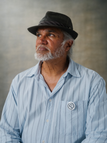A middle-aged man with a gray beard and a fedora wearing a light blue striped button-down shirt with a circular logo pin on the pocket.