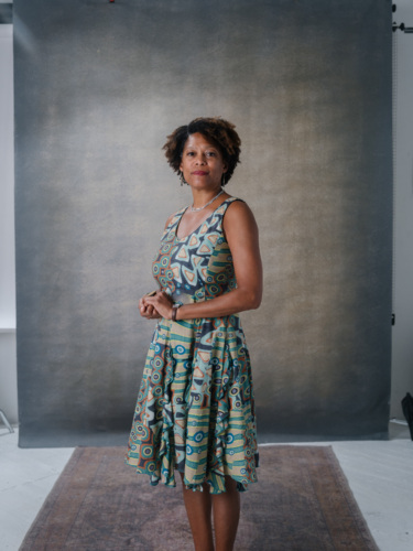A Black woman with short curly hair wearing a sleeveless patterned dress and a necklace, standing in front of a gray backdrop.