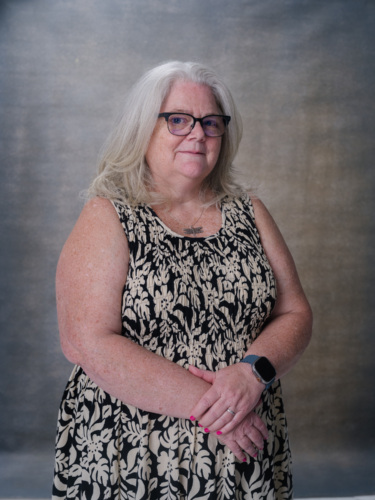 A woman with shoulder-length gray hair and glasses wearing a black and white patterned sleeveless dress.