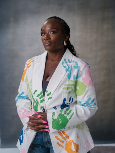 A woman with braided hair wears a white blazer patterned with colorful handprints.