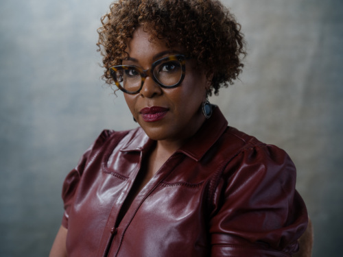 Portrait of a woman with curly hair wearing glasses and a maroon collared top.