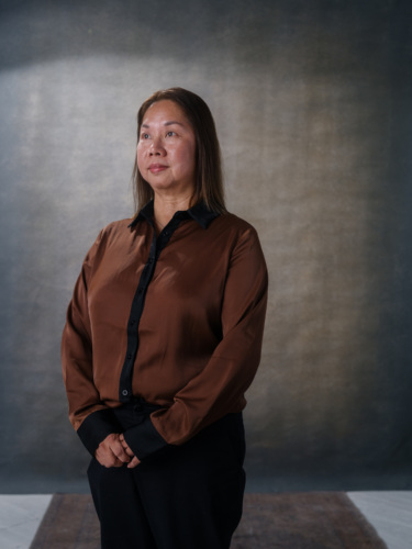 A middle-aged woman with shoulder-length hair wearing a brown button-up shirt and black trousers, standing against a dark backdrop.