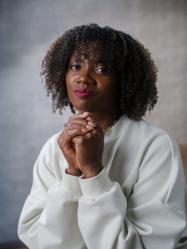 A woman with curly hair wearing a white sweatshirt, posing with her hands clasped under her chin.