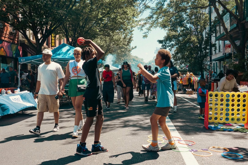 Two children play a game with plastic hoops and a ball on a street during an outdoor event, with pedestrians walking in the background.
