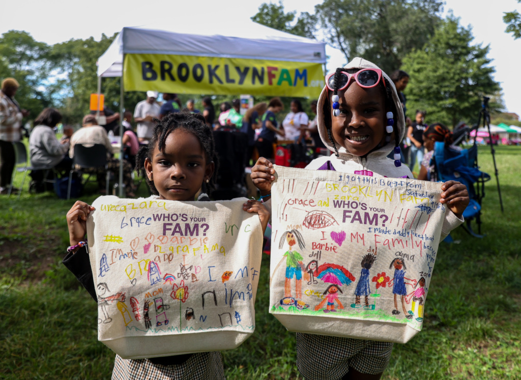 Two young girls holding up canvas tote bags decorated with hand-drawn pictures and text at an outdoor event with a BrooklynFam banner in the background.