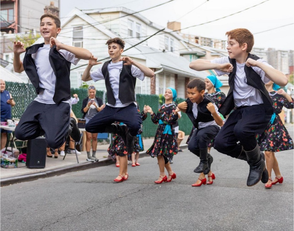 A group of children in traditional clothing performing a synchronized jump during an outdoor street dance.