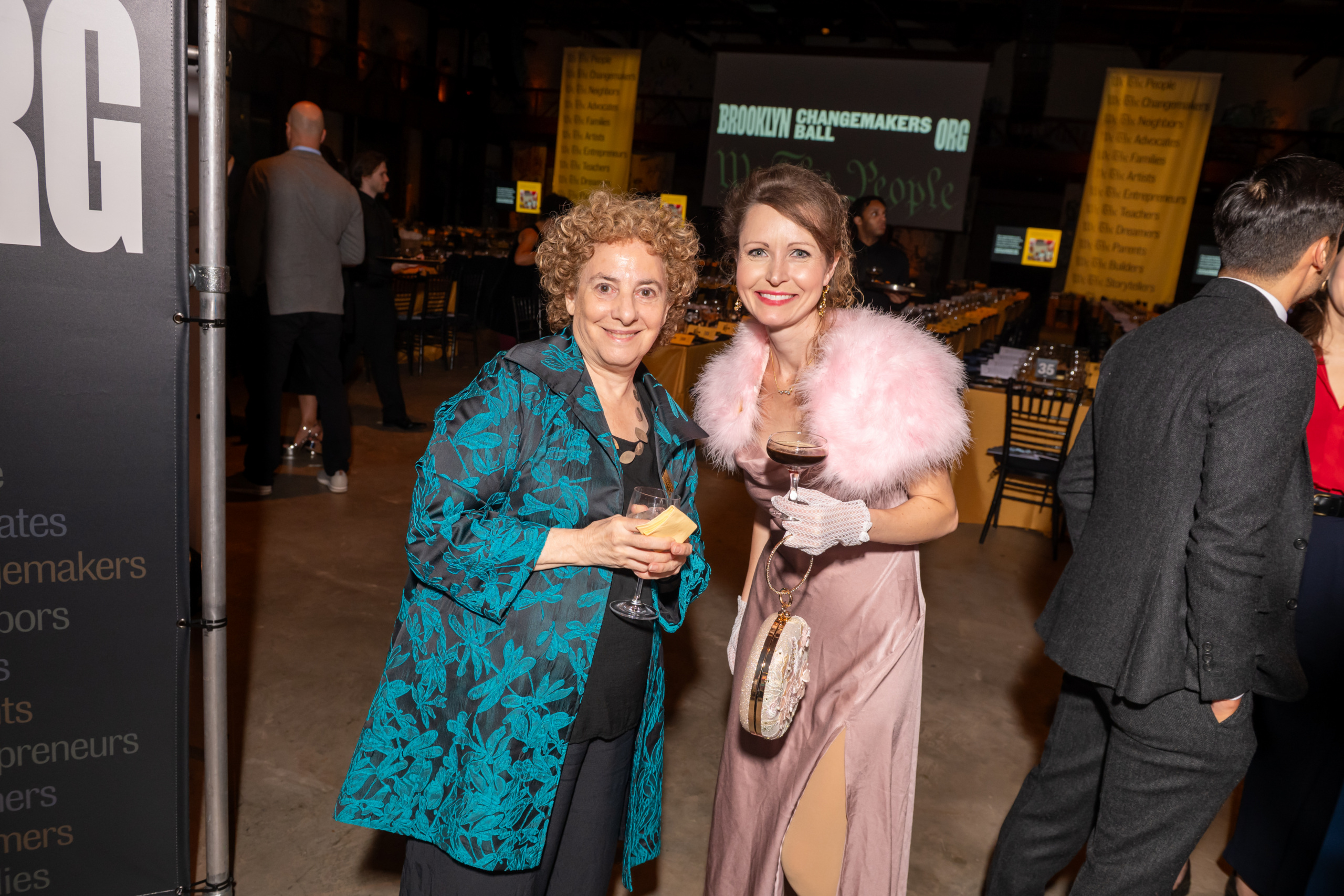 Two women smiling at the Brooklyn Changemakers Ball, one holding a glass of wine and the other holding a small plate.