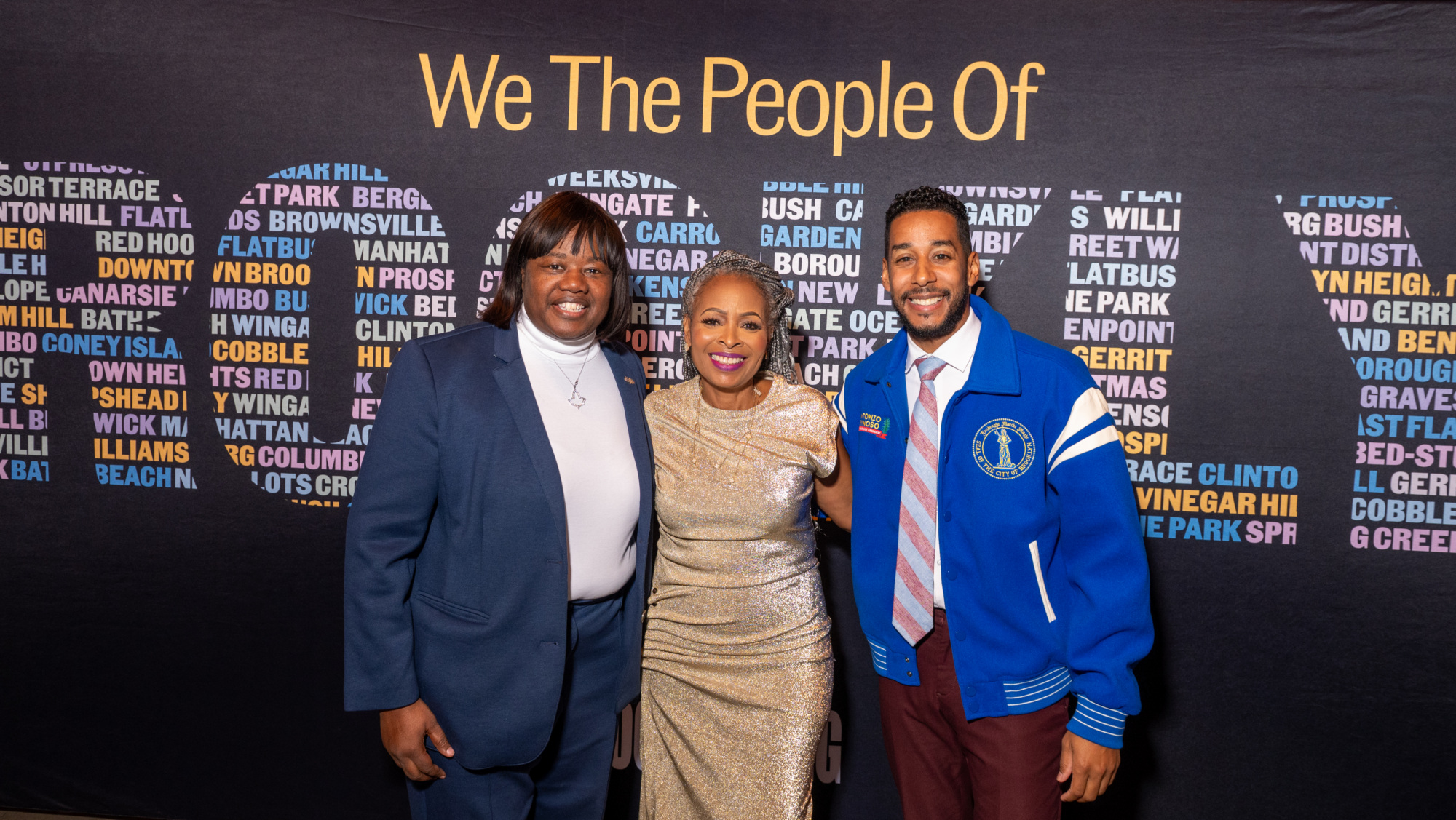 Three people stand in front of a black backdrop with the text “We The People Of” above a list of neighborhood names.