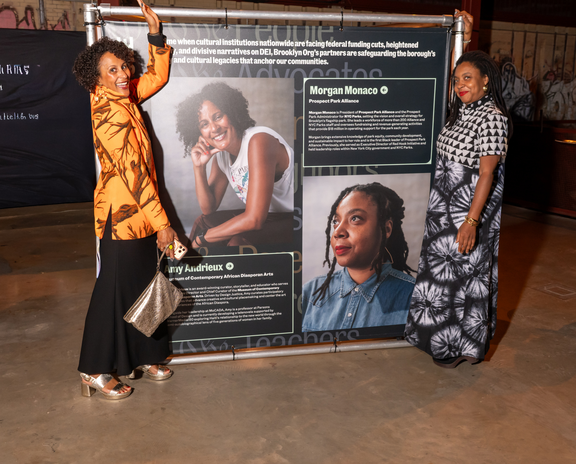 Two women stand beside a large exhibition banner featuring portraits and biographies of Morgan Monaco and Amy Andrieux.