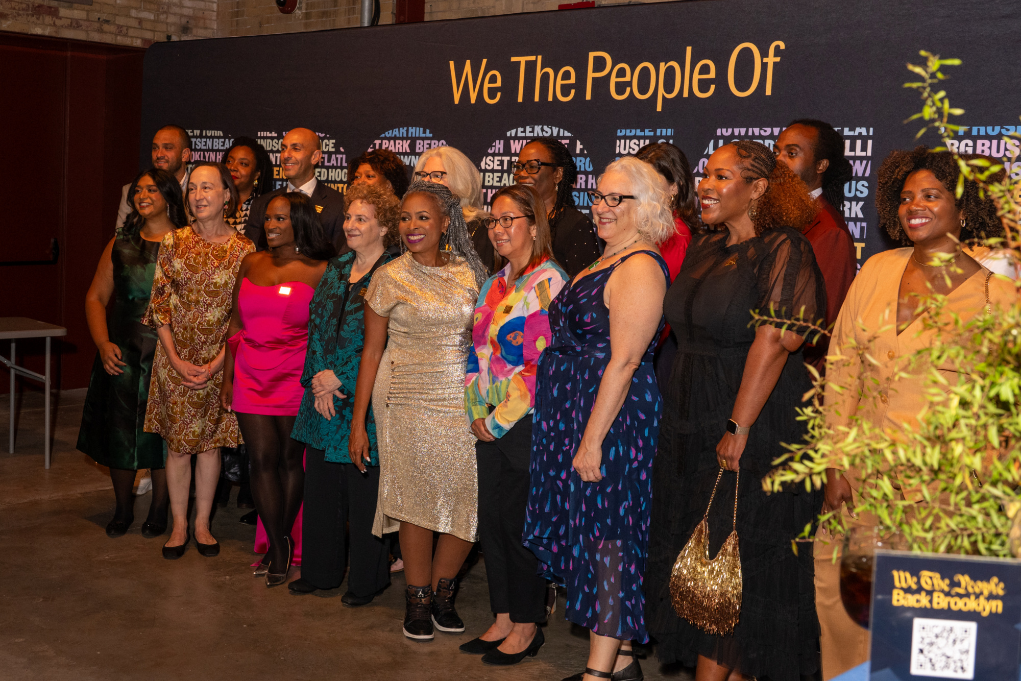 A diverse group of women standing in front of a black backdrop that reads “We The People Of” with a list of Brooklyn neighborhoods.