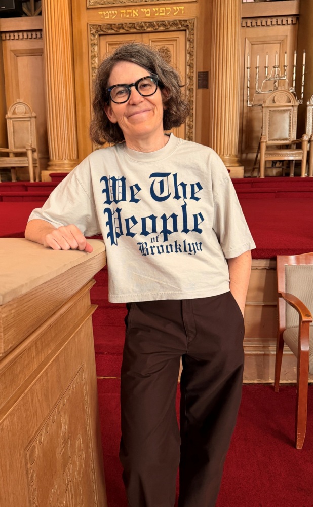 A person with glasses, wearing a shirt that says “We The People of Brooklyn,” standing in a synagogue.
