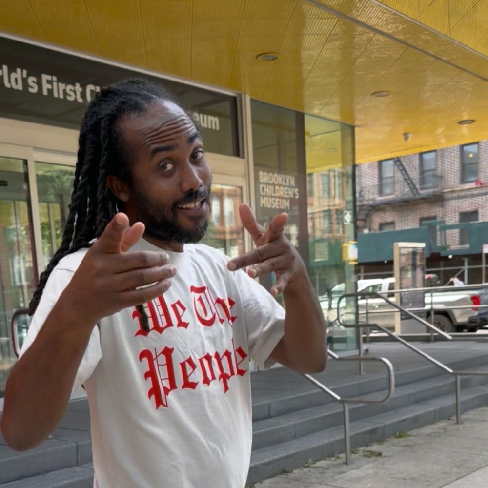 A person with locs wearing a “We The People” t-shirt points both hands toward the camera in front of the Brooklyn Children’s Museum.