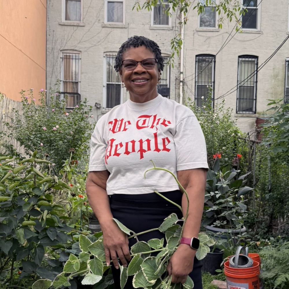 A smiling woman wearing a “We The People” t-shirt holds a green plant in a garden.