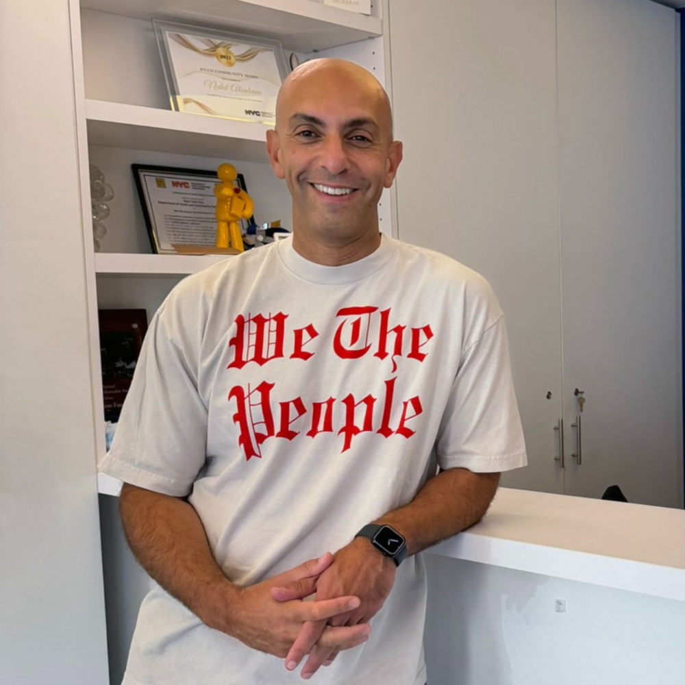 A smiling man wearing a white t-shirt with the words “We The People” in red gothic lettering, leaning against a white counter.